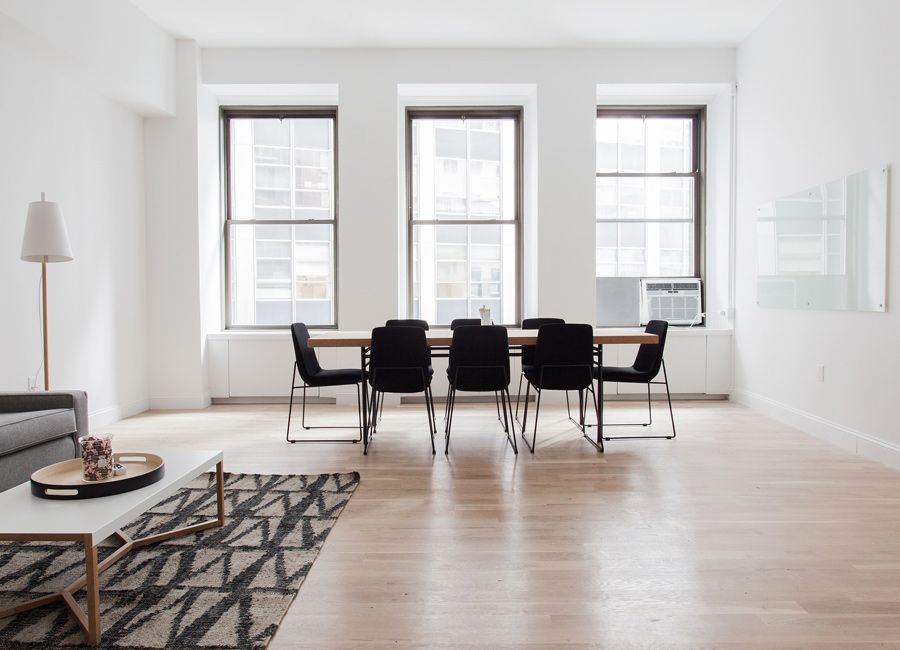 The image showcases a bright, spacious dining area with large windows allowing natural light to flood in. A long wooden dining table is surrounded by black chairs. A minimalistic coffee table sits in the foreground with a patterned rug beneath it. The walls are painted in a soft white, providing a clean and modern aesthetic.