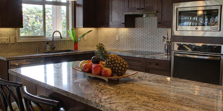 A modern kitchen featuring a stylish granite countertop with a central fruit bowl containing apples and a pineapple. The cabinetry is dark wood, and there are stainless steel appliances, including an oven and a microwave. The kitchen is well-lit by natural light streaming in through a window that overlooks greenery outside. The backsplash consists of small, light tiles that complement the countertop.