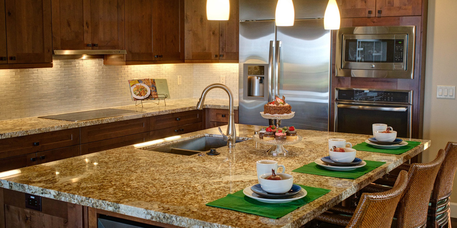 The image showcases a modern kitchen featuring a large island with a granite countertop. The kitchen cabinetry is made of rich, dark wood, and there are stainless steel appliances including a refrigerator and microwave. Pendant lights hang above the island, illuminating the space. A stylish cake stand displays desserts, and place settings with bowls are arranged on a green table runner. The backsplash consists of light tiles, adding a sleek touch to the overall design.