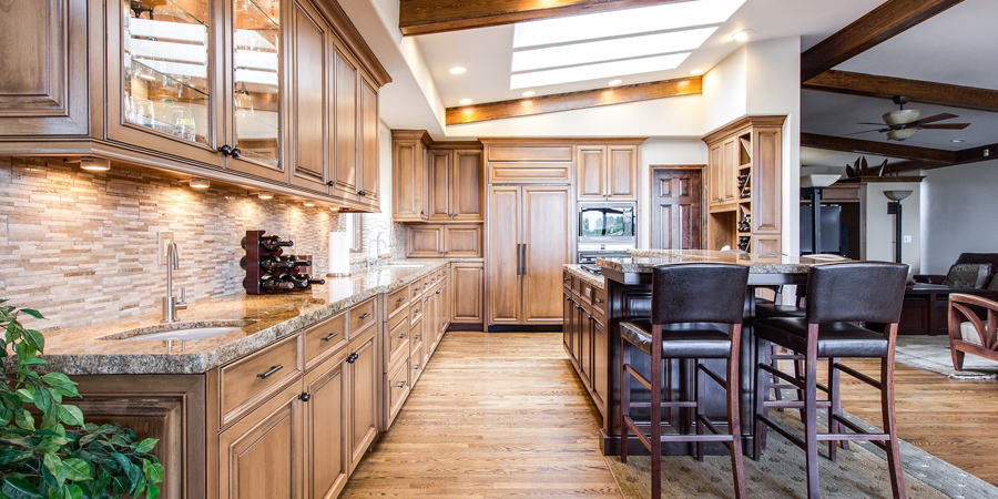 The image showcases a modern kitchen featuring rich wooden cabinetry and granite countertops. The kitchen is well-lit with natural light streaming in from a skylight. The floor is made of warm-toned hardwood, complementing the cabinetry. A kitchen island with seating is positioned in the centre, and the space is decorated with subtle, stylish accents.